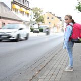 Eine Schülerin steht auf einem Gehweg neben einer Straße, über die ein Auto fährt.